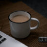 Eine weiße Kaffetasse, gefüllt mit Kaffe, steht neben einem Laptop auf einer braunen Tischunterlage.