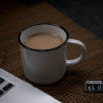 Eine weiße Kaffetasse, gefüllt mit Kaffe, steht neben einem Laptop auf einer braunen Tischunterlage.