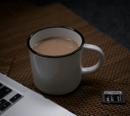 Eine weiße Kaffetasse, gefüllt mit Kaffe, steht neben einem Laptop auf einer braunen Tischunterlage.