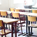 Ein heller Klassenraum mit den typischen, Holzstühlen und Tischen einer Schule bestückt.